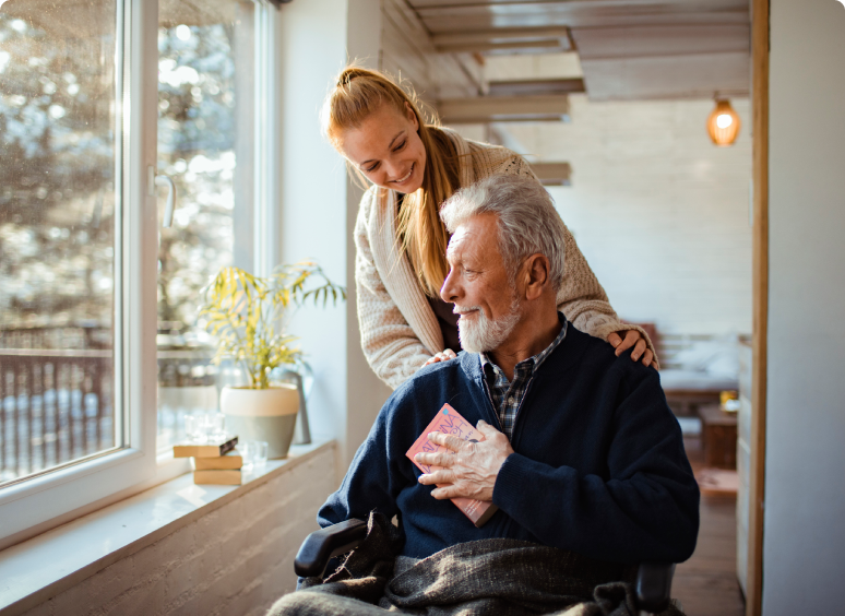 A young woman lovingly embraces an elderly man in a wheelchair by a window.