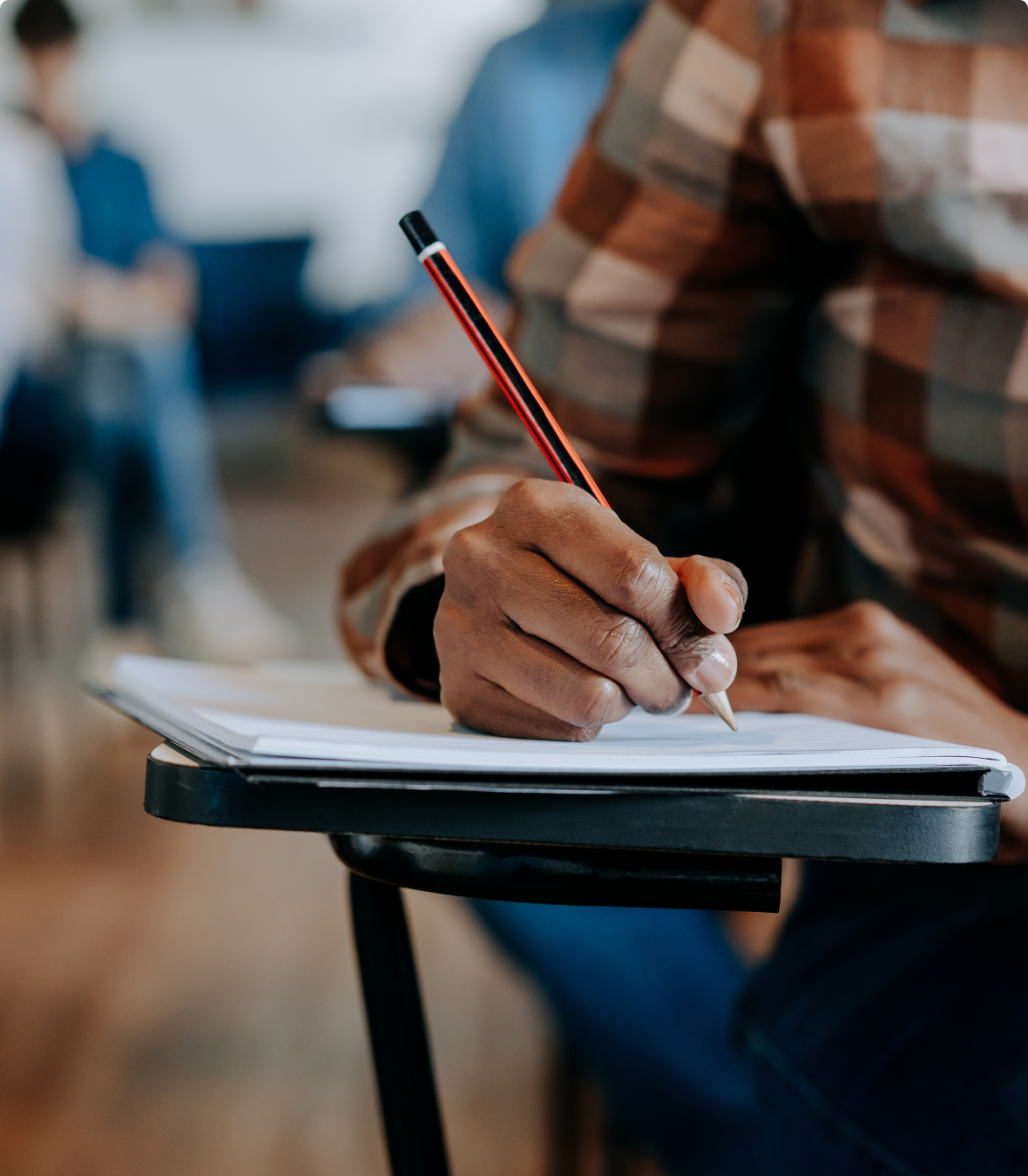 Person writing with a pencil on paper in a classroom.