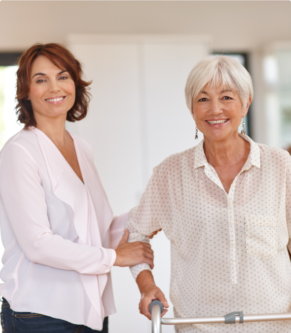 A caregiver supports an elderly woman with a walker indoors.